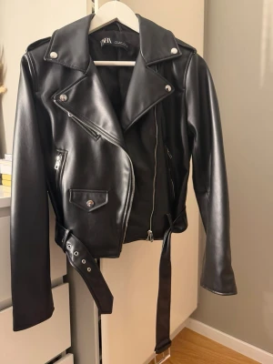 Svart  skinnjacka från Zara - Snygg svart skinnjacka från Zara i klassisk bikerstil med breda slag, silverfärgade dragkedjor och knappar samt bälte i midjan. Den är använd 1 gång, Är i storlek M men passar även mig som har S. Jackan har flera fickor och axelklaffar för en cool look. Perfekt för dig som vill ha en tidlös och edgy stil.