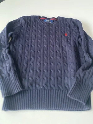 Mörkblå kabelstickad tröja Polo Ralph Lauren M 10-12 - Kabelstickad mörkblå tröja från Polo Ralph Lauren med rund halsringning och röd broderad logga på bröstet. Tröjan har långa ärmar och breda ribbade muddar vid ärmslut och nederkant. Perfekt för lager på lager och klassisk stil. 