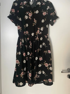 Svart blommig kortklänning - Svart kortklänning med blommigt mönster i vitt och rosa. Klänningen har korta ärmar, krage och knappar framtill. Perfekt för en somrig look och har en avslappnad passform med markerad midja.