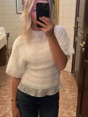 Vit blus med puffärm från Nelly - Supersöt vit blus från Nelly med puffiga korta ärmar och smockad struktur. Blusen har en volang nertill och är tillverkad i ett lätt och luftigt tyg som känns perfekt för varmare dagar.