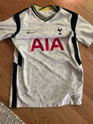 Tottenham Hotspur matchtröja Nike - Snygg Tottenham Hotspur matchtröja från Nike i vitt med mörkblå och gula detaljer. Tröjan har klubbmärke, Nike-logga och AIA sponsortryck på bröstet. Tillverkad i lätt och ventilerande material, perfekt för fotbollsträning eller match.