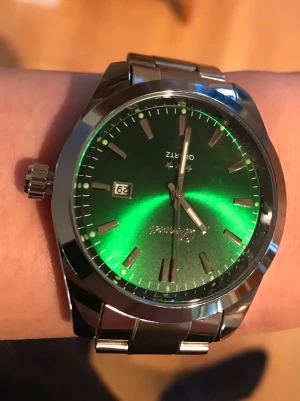 grön klocka i metall - Stilren klocka från Zanvo med metallarmband och en riktigt snygg grön urtavla. Klockan har datumvisning och silverfärgade detaljer på urtavlan. Perfekt accessoar för dig som vill sticka ut med en unik färg.