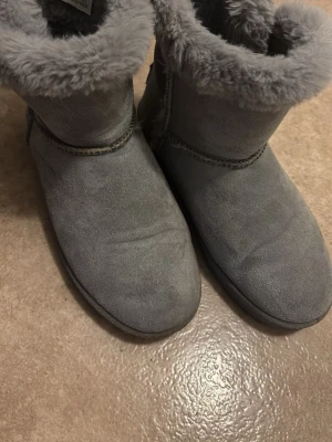 Gråa fluffiga vinterstövlar - Mysiga gråa stövlar med fluffig fuskpäls runt skaftet och mjuk mockaliknande yta. Klassisk rund tå och mellanhögt skaft, perfekta för kalla dagar. Snygg söm längs kanten ger extra detalj.