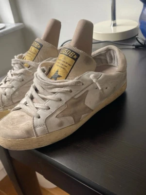 Beige sneakers från Golden Goose -  Stora i storleken Snygga beige sneakers från Golden Goose med den ikoniska stjärnan på sidan. Skorna har en vintage-look, vita snören och beige sula. Tillverkade i mocka och läder för en cool och avslappnad stil.
