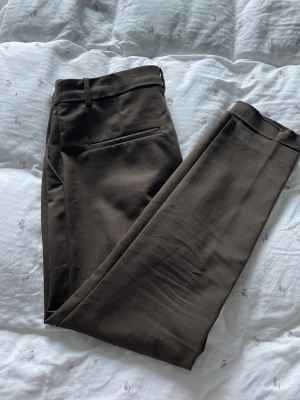 Bruna kostymbyxor med raka ben - Snygga bruna kostymbyxor med raka ben och pressveck. Byxorna har hällor för skärp, dold dragkedja och två bakfickor. Perfekta för en clean och stilren look. Materialet känns mjukt och har en lätt stretch.