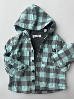 Rutig overshirt med huva från Zara - Mintgrön och grå rutig overshirt från Zara med huva och knäppning framtill. Två bröstfickor, varav en med patch där det står 'Walk the Route'. Mjuk insida och långa ärmar med knapp vid ärmslut. Perfekt för lager på lager.