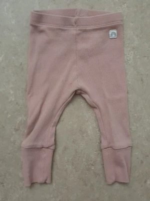 Leggings - Säljer ett par mjuka, ribbade leggings i ljusrosa färg med breda muddar och uppvik vid bensluten. De har en liten patch med regnbågsmotiv vid linningen och är perfekta för en avslappnad och trendig look.
