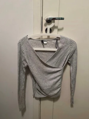 Ljusgrå omlottopp med ribbad struktur - Snygg ljusgrå omlottopp med ribbad struktur och v-ringad hals. Toppen har långa ärmar och sitter figurnära, perfekt för att styla med jeans eller kjol. Materialet känns mjukt och stretchigt, vilket gör den bekväm att bära hela dagen.