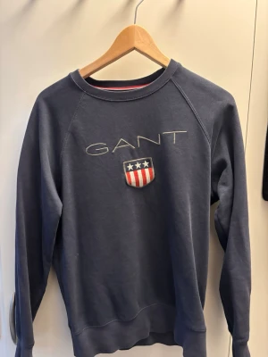 Blå sweatshirt från GANT - Mörkblå sweatshirt från GANT med broderad logga och sköld på bröstet. Tröjan har rund halsringning, ribbade muddar och klassisk collegekänsla. Perfekt för en avslappnad och stilren look.