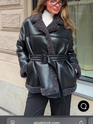 Svart skinnjacka med bälte Gina Tricot - Snygg svart skinnjacka från Gina Tricot med markerat bälte i midjan och grå fuskpälsdetaljer vid krage, ärmslut och nederkant. Jackan har en oversized passform och bred krage som ger en cool look.