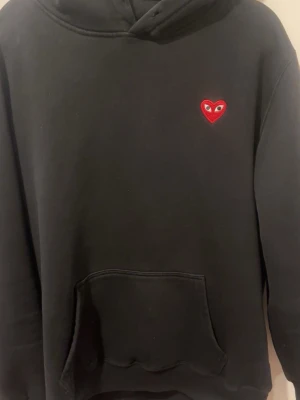 Svart hoodie med rött hjärta - Svart hoodie från Play med det ikoniska röda hjärtat med ögon broderat på bröstet. Stor ficka framtill och huva. På ryggen finns en rektangulär grafisk print i rosa och beige toner. Perfekt för dig som vill sticka ut lite. Storleken står de att de är xxl men jag är 180 och den passar perfekt, så skulle säga att den sitter som M