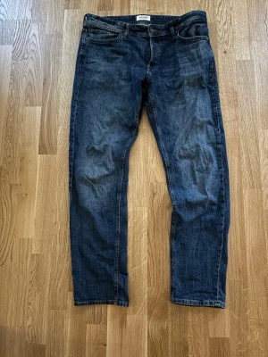 Mörkblå raka jeans från Jack & Jones  - Säljer ett par mörkblå jeans från Jack & Jones med klassisk rak passform. Jeansen har fem fickor, och en snygg tvättad look. Tillverkade i bomull med en skön känsla och stilren design. Perfekta till vardagslooken!