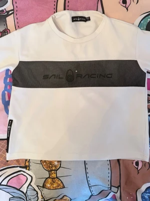 Vit t-shirt från Sail Racing - Vit t-shirt från Sail Racing med svart panel och logga över bröstet. Klassisk rund halsringning och korta ärmar. Tillverkad i mjuk bomull som känns skön mot huden. Perfekt för en clean och sportig look.