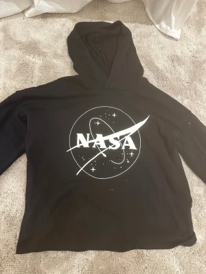 Svart NASA hoodie med tryck - Cool svart hoodie med stor NASA-logga och rymdmotiv i vitt på bröstet. Hoodien har en klassisk huva och är gjord i mjukt material, perfekt för dig som gillar rymd och streetwear. Passar till jeans eller joggers för en avslappnad look. Den har några fläckar men går säkert att tvätta bort.