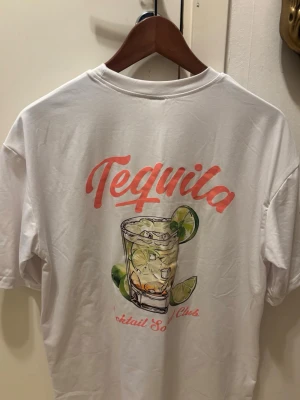 Vit t-shirt med tequila-tryck - Vit t-shirt i bomull med färgglatt tryck av ett tequila-glas och lime på både bröstet och ryggen. Texten 'Tequila' i rosa och orange ger en chill vibe. Klassisk passform och rund halsringning, perfekt för sommarens alla äventyr.