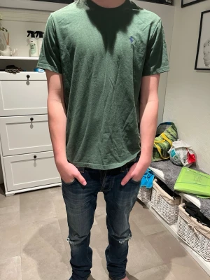 Grön t-shirt från Polo Ralph Lauren - Mörkgrön t-shirt från Polo Ralph Lauren med klassisk rund halsringning och korta ärmar. T-shirten har den ikoniska broderade loggan i marinblått på bröstet. Tillverkad i mjuk bomull för en skön känsla och enkel stil.