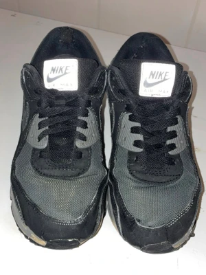 Nike Air Max svarta sneakers - Klassiska Nike Air Max sneakers i svart och grått med mesh och syntetmaterial. Swoosh-logga på sidan och tydlig Air Max-dämpning i sulan. Skorna har snörning och en sportig, chunky siluett som passar till streetwear.