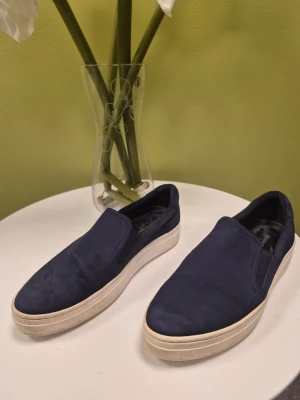 Vagabond mörkblåa slip-on suede loafers 37 - Säljer ett par mörkblåa slip-on loafers med vit sula. Skorna har en stilren och enkel design utan snörning, vilket gör dem lätta att ta på. Ovandelen är i mockaliknande material och sulan är i gummi. Perfekta för en avslappnad och trendig look.