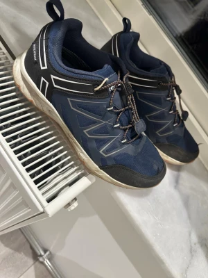 Blå sneakers från Salomon - Säljer ett par blå sneakers från Salomon med svarta och vita detaljer. Skorna har snabbsnörning, slitstark ovandel i textil och syntet samt förstärkt tåparti. Yttersulan är vit med orange detaljer undertill. Perfekta för en aktiv livsstil.