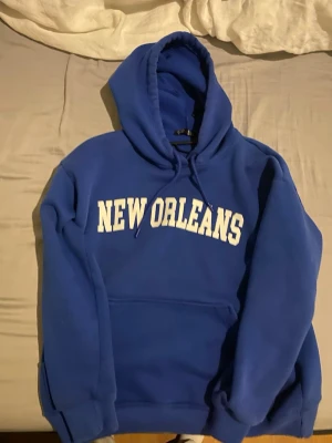 Blå hoodie med New Orleans-tryck - Säljer en blå hoodie med vit text 'NEW ORLEANS' på bröstet. Hoodien har en stor huva, dragsko och en klassisk magficka. Perfekt för dig som gillar college-stil och vill ha något bekvämt och snyggt. Materialet känns mjukt och tjockt.