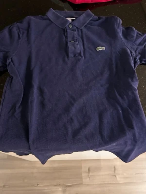 Mörkblå pikétröja från Lacoste - Klassisk mörkblå pikétröja från Lacoste med den ikoniska broderade krokodilloggan på bröstet. Tröjan har korta ärmar, krage och knappar framtill. Tillverkad i bomull för en bekväm och luftig känsla.