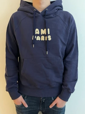 Mörkblå hoodie från AMI Paris - Snygg mörkblå hoodie från AMI Paris med broderad text i vitt på bröstet. Klassisk känguruficka framtill, justerbar huva med dragsko och ribbade muddar. Perfekt för en avslappnad och trendig look.