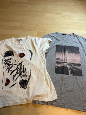 2-pack t-shirts med tryck - Säljer två t-shirts i storlek S. Ena är vit med ett stort sneakers-tryck i svart och rött, den andra är grå med ett grafiskt tryck av en vägskylt och text. Båda har rund hals och korta ärmar, perfekta till vardags. Materialet känns mjukt och skönt, troligtvis bomull.