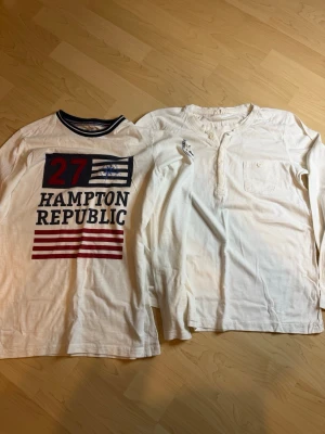 2 vita långärmade tröjor Hampton Republic - Två vita långärmade tröjor från Hampton Republic. Ena tröjan har tryck med texten '27 Hampton Republic' och marinblå detaljer vid kragen. Den andra är enkel med knappar vid halsen och bröstficka. 
