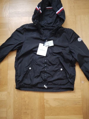 Svart vindjacka från Moncler - Snygg svart vindjacka från Moncler med huva och dragkedja framtill. Jackan har vita och röda ränder på huvan, två fickor med tryckknappar och Moncler-logga på ärmen. Tillverkad i lätt polyester som passar perfekt för blåsiga dagar.