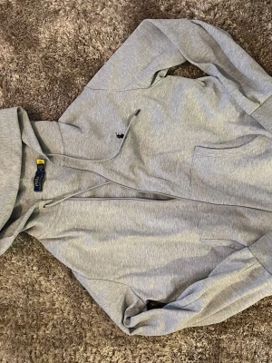 Grå hoodie från Polo Ralph Lauren - Snygg grå hoodie från Polo Ralph Lauren med dragkedja framtill och klassisk huva med dragsko. Broderad logga i svart på bröstet och känguruficka. Tillverkad i mjukt bomullsmaterial som är perfekt för chill dagar.