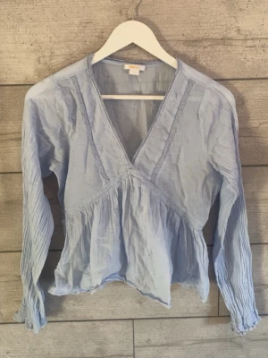 Ljusblå v-ringad blus med volang - Supersöt ljusblå blus med v-ringning och volang nedtill. Blusen har långa ärmar med smock vid ärmsluten och tunna dekorativa sömmar framtill. Tillverkad i ett lätt och luftigt tyg som ger en avslappnad känsla.