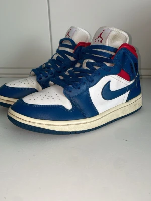 Nike Air Jordan 1 Mid blå/vit/röd - Säljer ett par Nike Air Jordan 1 Mid sneakers i blått, vitt och rött. Skorna har klassisk swoosh på sidan, perforerad tåbox och högt skaft. Tillverkade i läder med textildetaljer och har blå snören. Perfekt för dig som gillar retrostil och streetwear.