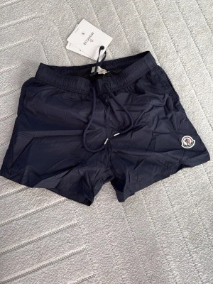 Mörkblå badbyxor från Moncler - Snygga mörkblå badbyxor från Moncler med klassisk logotyp på vänster ben. Modellen har elastisk midja med dragsko och innerbyxa i mesh. Tillverkade i lätt och snabbtorkande material, perfekta för stranden eller poolen.