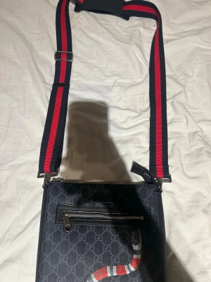 Gucci axelväska med orm och GG-mönster - Snygg axelväska från Gucci i svart canvas med klassiskt GG-mönster och en röd, vit och svart orm på framsidan. Väskan har en justerbar axelrem i rött och marinblått, samt en praktisk ytterficka med dragkedja. Perfekt för dig som vill sticka ut.