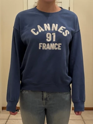 Blå sweatshirt med tryck - Blå sweatshirt med vit text 'CANNES 91 FRANCE' på bröstet. Tröjan har rund halsringning, långa ärmar och ribbade muddar vid ärmslut och nederkant. Perfekt för en avslappnad och cool stil.