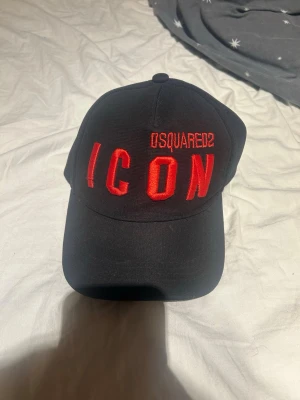 Svart Dsquared2 ICON keps - Säljer en svart keps från Dsquared2 med stor röd brodyrtext 'ICON' och Dsquared2-logga framtill. Klassisk böjd skärm och justerbar rem bak. Tillverkad i bomull för en bekväm och snygg look.