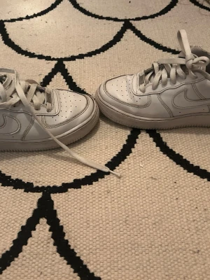 Vita Nike Air Force 1 sneakers - Klassiska vita Nike Air Force 1 sneakers i lågt utförande. Skorna har en stilren design med perforerad tå, vita skosnören och den ikoniska swoosh-loggan på sidan. Tillverkade i läder med en robust gummisula för extra komfort och streetkänsla.