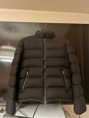 Svart pufferjacka från Herno - Snygg svart pufferjacka från Herno med hög krage och två dragkedjefickor framtill. Jackan har en stilren design med breda, vadderade paneler och dragkedja hela vägen upp. Perfekt för kalla vinterdagar och riktigt trendig look.