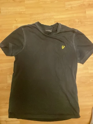 Svart t-shirt från Lyle & Scott - En svart t-shirt från Lyle & Scott med klassisk passform och korta ärmar. T-shirten har en gul broderad logga på bröstet och är tillverkad i mjuk bomull. Perfekt till vardags för en clean och enkel stil.
