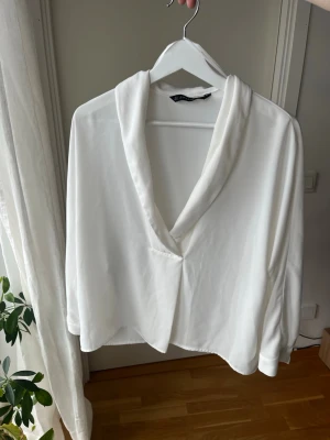 Vit blus med djup urringning från Zara - Stilren vit blus från Zara med snygg sjalkrage och avslappnad passform. Blusen har långa ärmar och är tillverkad i ett mjukt, lätt material som ger en elegant känsla. Perfekt att matcha med jeans eller kjol för en trendig look.
