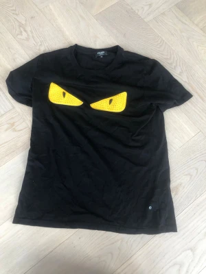 Svart Fendi t-shirt med gula ögon - Cool svart t-shirt från Fendi med ikoniska gula ögon framtill. Tillverkad i mjuk bomull och har en klassisk passform med korta ärmar och rund hals. Perfekt för dig som vill sticka ut med en unik designertee.
