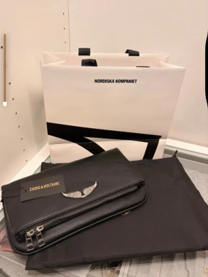 Zadig & Voltaire Väska - Snygg svart handväska från Zadig & Voltaire i äkta läder. Väskan har en silverfärgad vinge framtill och dubbla dragkedjor. Kommer med original dustbag och tag. Aldrig använd, helt ny fick i present!