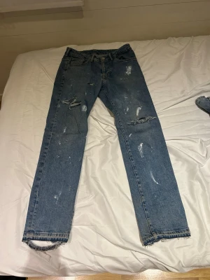 Blå slitna raka jeans - Säljer ett par blå jeans med rak passform och slitna detaljer, inklusive revor och fransade benslut. Jeansen har klassisk femficksdesign och är tillverkade i denim. Perfekta för en avslappnad och trendig look.