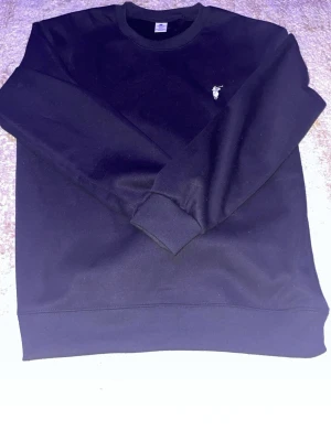 Svart sweatshirt med liten logga - En stilren svart sweatshirt med rund hals och liten broderad logga på bröstet. Tröjan har ribbade muddar vid ärmslut och nederkant, perfekt för en avslappnad och trendig look. Tillverkad i mjukt material som känns skönt mot huden.