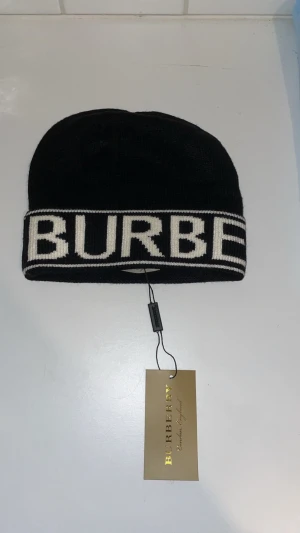 Svart mössa från Burberry - Snygg svart mössa från Burberry med bred vit kant och stor logotyp i vitt runt hela mössan. Klassisk beanie-modell i mjukt stickat material som ger en clean och modern look. Perfekt för dig som vill ha en stilren accessoar med lyxig känsla.