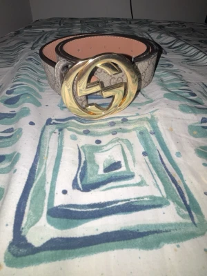 Gucci beige bälte med guldspänne  - Snyggt beige bälte från Gucci med klassiskt GG-mönster och stort guldigt spänne framtill. Bältet har bruna kanter och insidan är ljusrosa. Perfekt accessoar för att lyfta vilken outfit som helst.