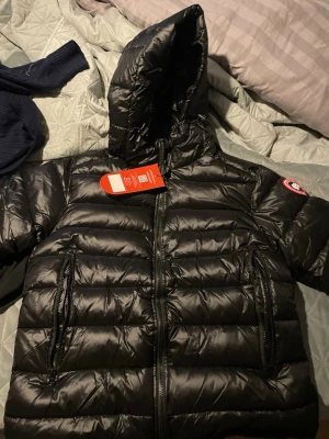 Svart dunjacka från Canada Goose - Snygg svart dunjacka från Canada Goose med huva och två stora dragkedjefickor framtill. Jackan har ett klassiskt quiltat mönster och ikonisk logga på ärmen. Perfekt för kalla vinterdagar och riktigt varm tack vare dunfyllningen.