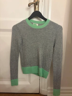 Grå tröja med gröna detaljer From Future - Snygg grå tröja från From Future med ribbstickade gröna muddar vid hals, ärmslut och nederkant. Tröjan är en lite kortare modell men sitter så snyggt på!. Perfekt för dig som gillar stilrena plagg med en twist. Använd ca 6 gånger, inte alls nopprig eller några hål💗