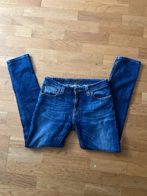Blå slim fit jeans med raka ben från nudie - Säljer ett par klassiska blå jeans i slim fit-modell med raka ben och fem fickor. Jeansen har en snygg tvättad look och kontrastsömmar. Perfekta för en avslappnad och trendig stil. Materialet är denim i bomull. (Nypris 2000 kr)