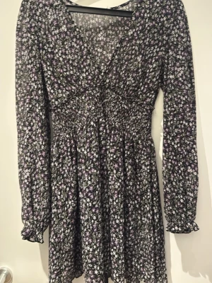 Blommig kortklänning med v-ringning - Söt kortklänning med svart botten och små vita och lila blommor. Klänningen har v-ringning, långa ärmar med resår vid ärmslut och smock i midjan för en smickrande passform. Perfekt för vår och sommar!